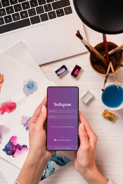 cropped view of designer at office desk holding smartphone with instgram app on screen, flat lay