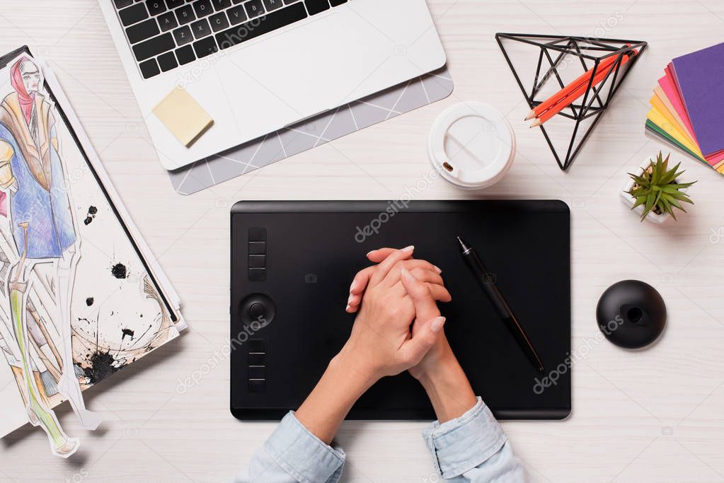 cropped view of designer holding hands above office desk with laptop, graphics tablet and pen