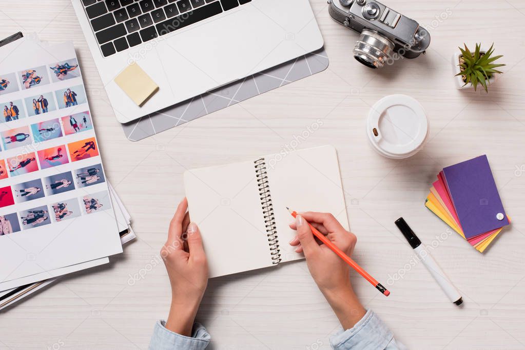 Cropped view of designer working at office desk and writing in notepad with laptop and designer supplies