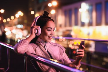 mutlu çekici Asyalı kız sokakta akşam, gelecekteki kavramının şehir içinde neon ışıklı smartphone ile müzik dinleme
