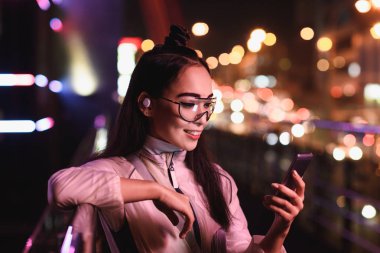 akşam, gelecekteki kavramının şehir sokak neon ışık ile smartphone kullanarak kablosuz kulaklık ile çekici Asyalı kız
