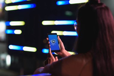 arkadan görünüşü akşamlar, gelecekteki kavramının şehir sokak neon ışık ile smartphone ile yüklenen shazam sayfa tutan kız