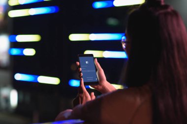 arkadan görünüşü akşamlar, gelecekteki kavramının şehir neon ışık ile sokakta smartphone ile yüklenen facebook sayfa tutan kız