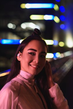 Kablosuz kulaklık uzakta akşamlar, gelecekteki kavramının şehir neon ışık ile sokakta görünümlü şık Asyalı kız gülümseyerek