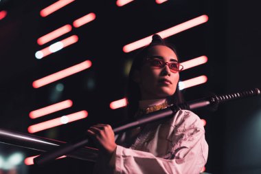 çekici Asyalı kız beyaz kimono holding Japon kılıcına düşük açılı görünüş Caddesi'nde akşam, gelecekteki kavramının şehir içinde neon ışıklı