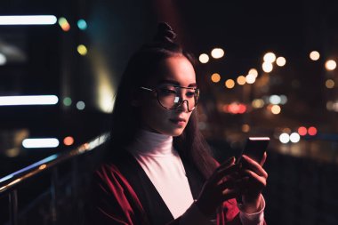 Bordo kimono ve gözlük akşamlar, gelecekteki kavramının şehir sokak neon ışık ile smartphone kullanarak çekici Asyalı kız