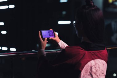 arkadan görünüşü cihaz neon ışık ile sokakta akşam, gelecekteki kavramının şehir alışveriş web ile smartphone tutan kız
