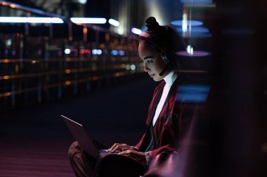 attractive asian girl in kimono sitting and using laptop with headphones on street with neon light, city of future concept clipart