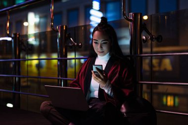 beautiful asian girl in kimono sitting and using laptop with smartphone on street with neon light, city of future concept clipart