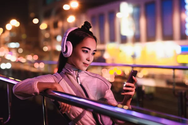 attractive asian girl listening music with smartphone on street with neon light in evening, city of future concept