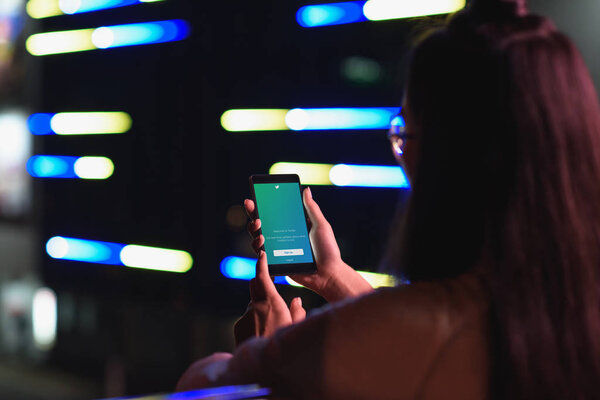 back view of girl holding smartphone with loaded twitter page on street with neon light in evening, city of future concept