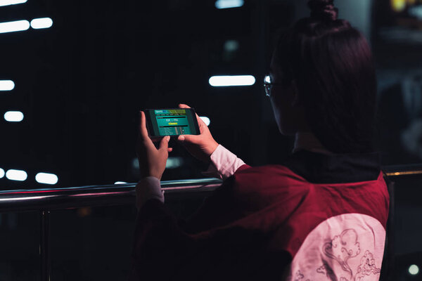 back view of girl holding smartphone with sports bet appliance on street with neon light in evening, city of future concept