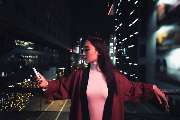 attractive asian girl leaning on railing and using smartphone on street with neon light in evening, city of future concept