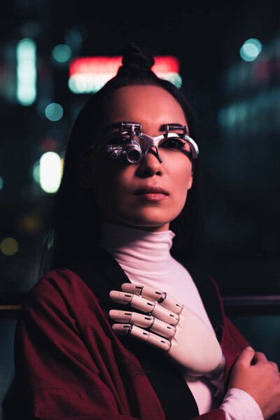 attractive asian girl with futuristic arm and eye prosthesis looking at camera on street in evening, city of future concept