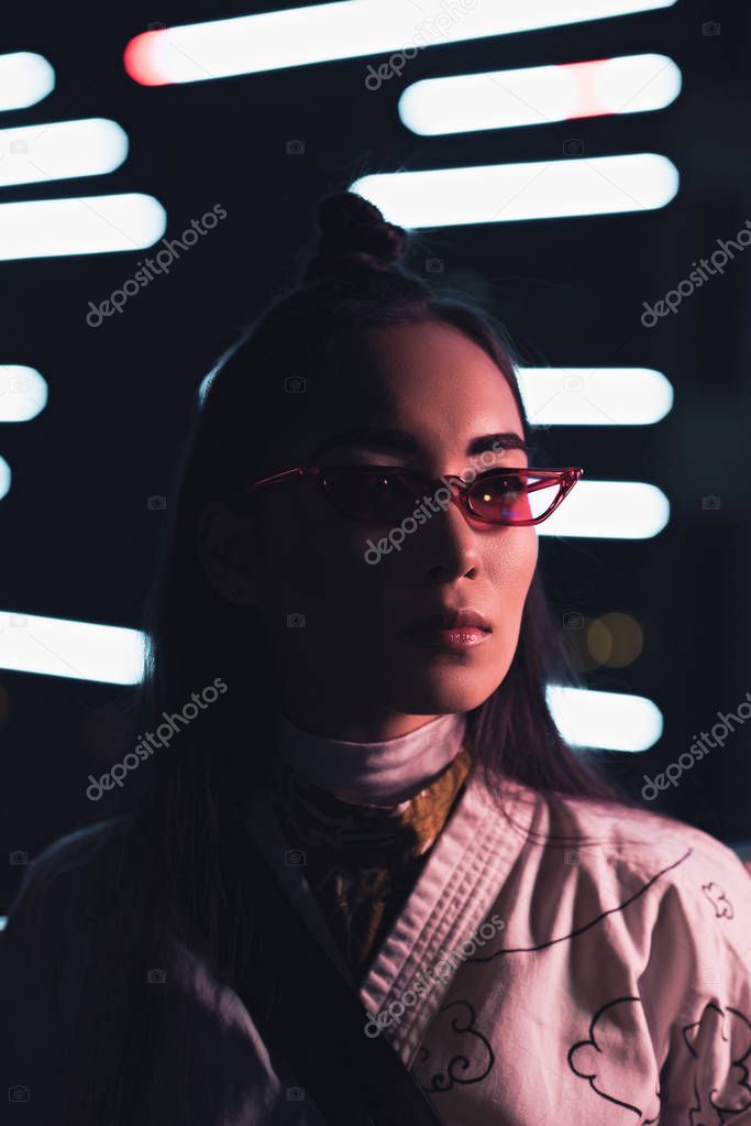 portrait of attractive asian girl in white kimono and sunglasses looking away on street with neon light in evening, city of future concept