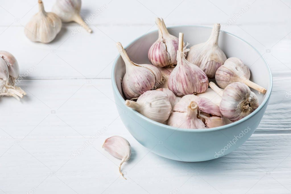 Blue ceramic bowl with heads of garlic and several peeled cloves on white wooden kitchen  table