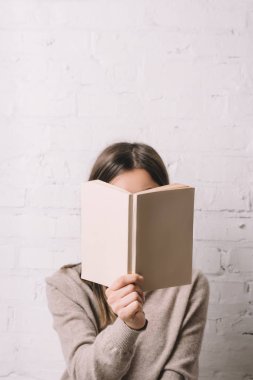 girl hiding face behind book near white brick wall clipart