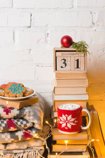 pile of books, blanket, gingerbread cookies, cup of hot chocolate with marshmallows, calendar with 31 december and christmas bauble