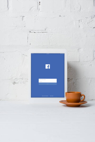 digital tablet with facebook application and cup of coffee near white brick wall 