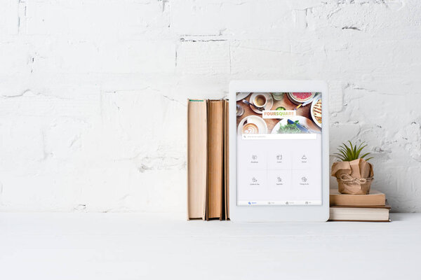 digital tablet with foursquare application, books and potted plant near white brick wall