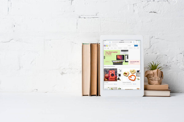 digital tablet with ebay application, books and green potted plant near white brick wall