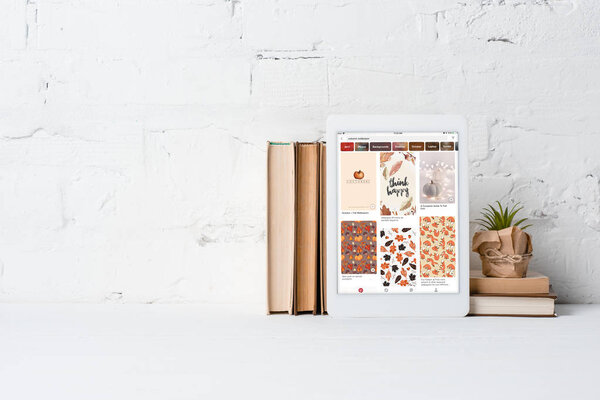 digital tablet with pinterest app, books and houseplant near white brick wall 