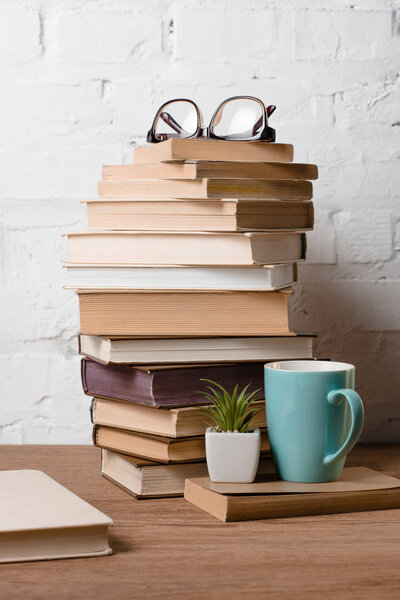 pile of books, eyeglasses, potted plant and cup with hot beverage on wooden table