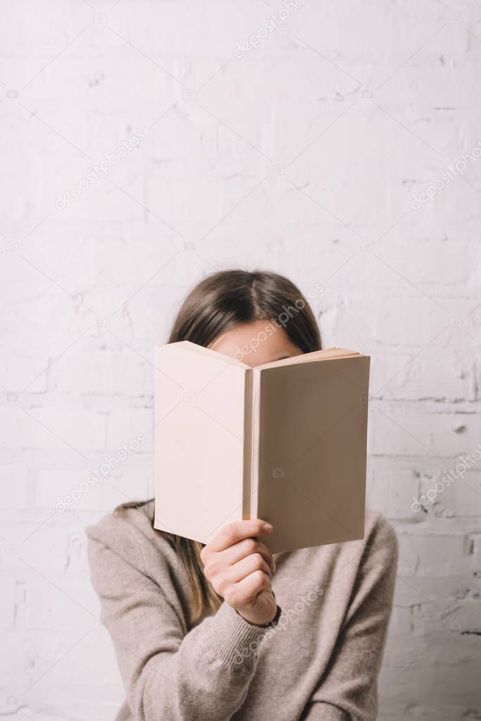 girl hiding face behind book near white brick wall