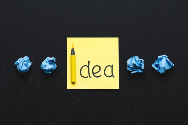 \'idea\' word written on sticky note and blue crumpled paper balls on black background, ideas concept