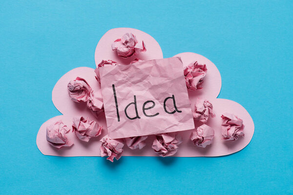 top view of pink cardboard cloud with crumpled paper balls and 'idea' word written on sticky note on blue background, ideas concept