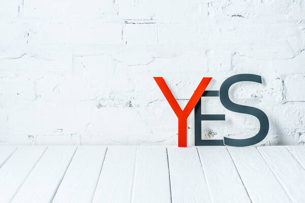 "yes" lettering with decorative red and black letters on wooden table near white brick wall
