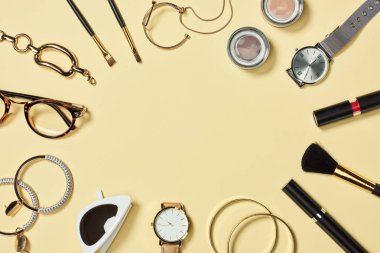 Top view of watches, lipstick, glasses, sunglasses, eyeshadow, blush, cosmetic brushes, bracelets, earrings and mascara on yellow background clipart