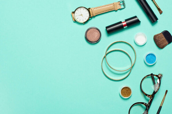 Top view of mascara, watch, lipstick, bracelets, eyeshadow, blush, glasses and cosmetic brushes with copy space
