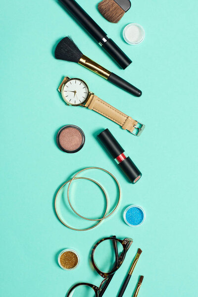 Mascara, watch, lipstick, bracelets, eyeshadow, blush, glasses and cosmetic brushes on turquoise background