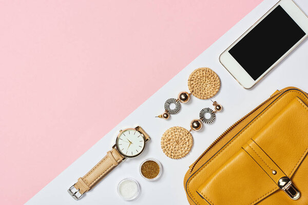 Top view of earrings, eyeshadow, watch, smartphone and yellow bag