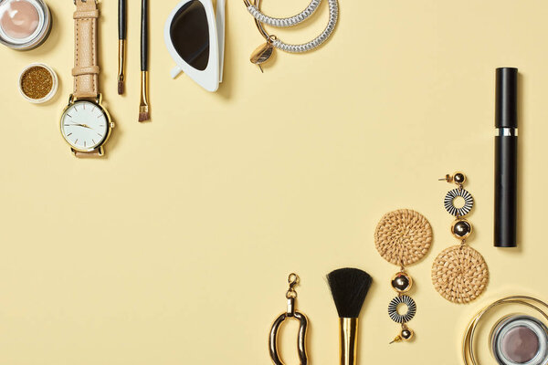 Top view of watch, sunglasses, eyeshadow, cosmetic brushes, bracelets, earrings and mascara on yellow background