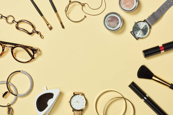 Top view of watches, lipstick, glasses, sunglasses, eyeshadow, blush, cosmetic brushes, bracelets, earrings and mascara on yellow background