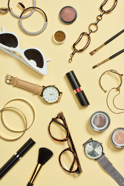 Top view of watches, lipstick, glasses, sunglasses, eyeshadow, blush, cosmetic brushes, bracelets, earrings and mascara 