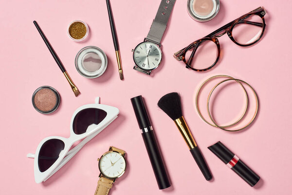 Top view of watches, lipstick, glasses, sunglasses, eyesshadow, blush, cosmetic brushes, bracelets and mascara
 