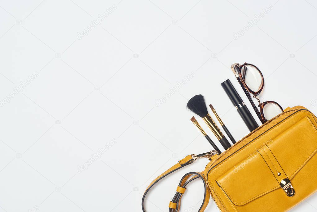 Top view of glasses, mascara, cosmetic brushes and yellow bag on white background