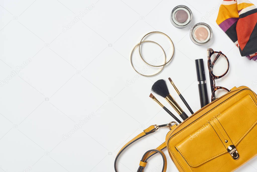 Top view of bracelets, scarf, glasses, mascara, cosmetic brushes, eyeshadow and yellow bag 