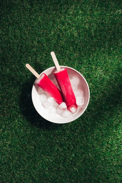 Tigela com Popsicles vermelhos e cubos de gelo no fundo de grama verde — Fotografia de Stock