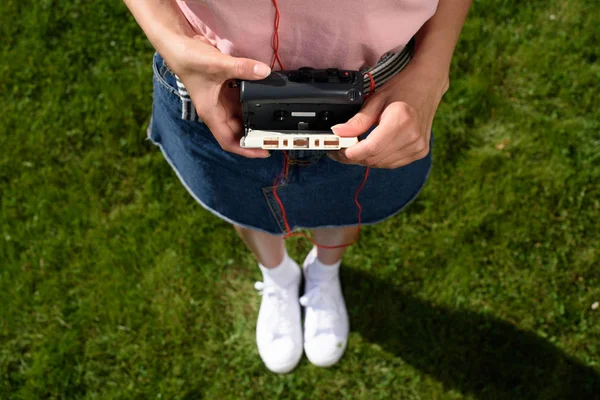Cropped shot of woman with earphones holding retro cassette player with audio cassette in hands while standing on green grass — Stock Photo