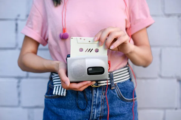 Cropped shot of woman with earphones holding retro cassette player and audio cassette in hands against white brick wall — Stock Photo