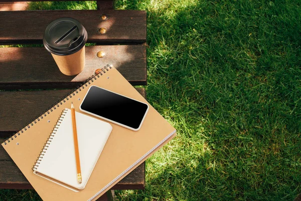 Vista de cerca del teléfono inteligente con pantalla en blanco, cuadernos y café para ir en banco de madera - foto de stock