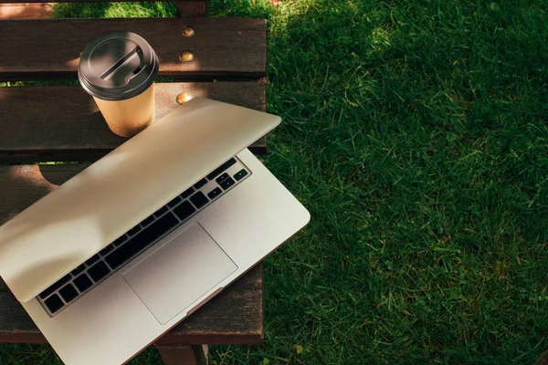 Vista de perto da xícara descartável de café e laptop no banco de madeira — Fotografia de Stock
