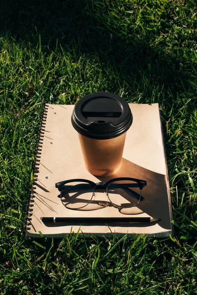 Vista de perto de café para ir, notebook com lápis e óculos na grama verde — Fotografia de Stock