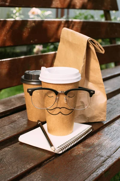 Vista de cerca de tazas desechables de café, bolsa de papel, anteojos y cuaderno en el banco - foto de stock