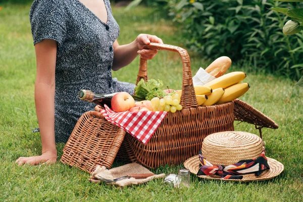 Imagen recortada de la mujer sentada sobre hierba verde en el picnic y tocando cesta con comida - foto de stock