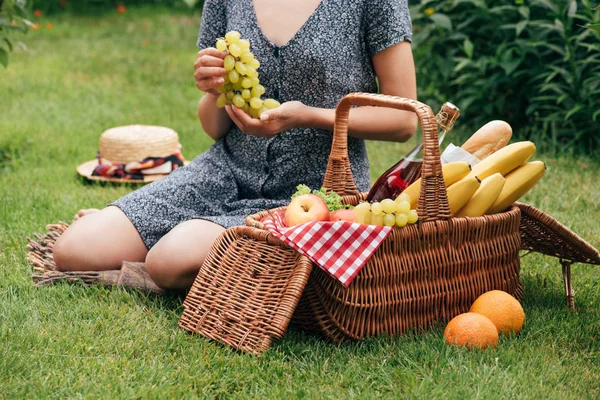 Обрезанный образ женщины, держащей виноград и сидящей на зеленой траве на пикнике — стоковое фото